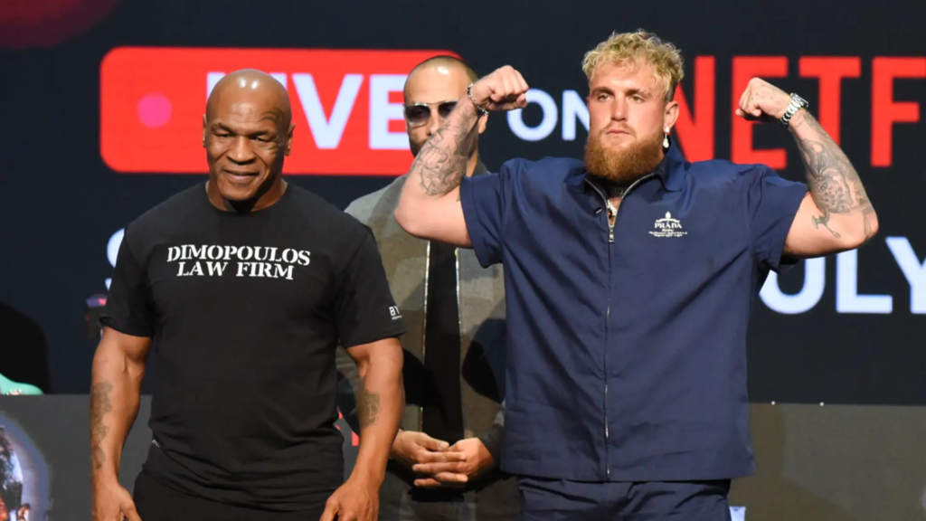 Jake Paul flexing his muscles alongside Mike Tyson at a promotional event, with Jake wearing a blue Prada outfit and Tyson in a black t-shirt with 'Dimopoulos Law Firm' text. The background shows a large screen with Netflix branding